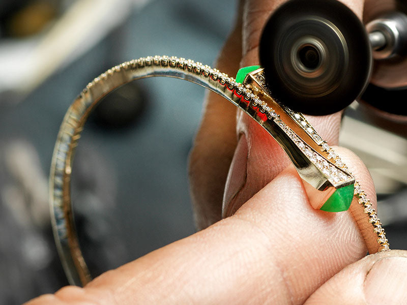 A close up of a person polishing a gold diamond bracelet with green stones with a polishing machine
