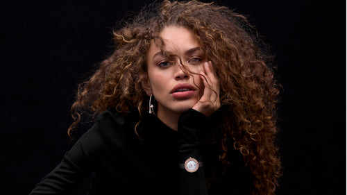 A close-up portrait of a woman with voluminous curly brown hair, wearing a black top and a round gold-toned pin or brooch on the chest, posed against a solid black background.