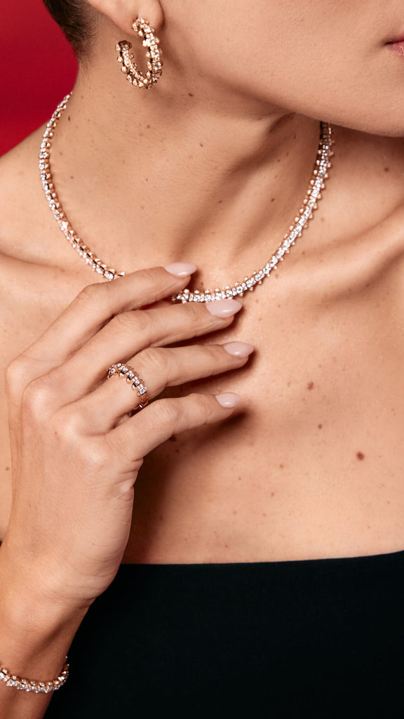 A close up of a woman's bare shoulders and hands, while she is touching a diamond necklace. She is wearing a sleeveless black top, with a diamond ring and earring in focus. 